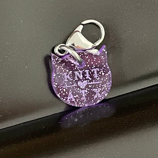 Purple glittery keychain with 'KNIT ATX' text, photographed on a dark surface.